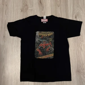 Svart Marvel Spiderman t-shirt - Cool svart t-shirt från Marvel med ett stort tryck av The Amazing Spiderman i serietidningsstil på framsidan. Perfekt för dig som gillar superhjältar och vill sticka ut med en ikonisk look.
