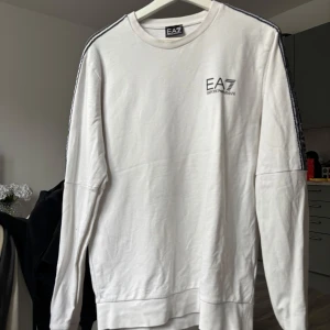 Vit sweatshirt från EA7 Emporio Armani - Säljer en vit sweatshirt från EA7 Emporio Armani med svarta detaljer längs ärmarna där loggan är tryckt. Tröjan har rund halsringning och långa ärmar. Perfekt för en stilren och sportig look.