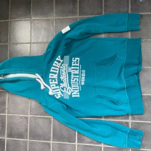 hoodie Superdry - Turkos hoodie från Superdry med snyggt tryckt logga och text på bröstet. Tjocka snören från luvan som ger en snygg detalj. Hoodien är i storlek M. Tveka inte skriva vid frågor!!!