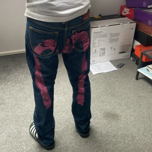 Evisu jeans med rosa målad detalj - Unika blå jeans från Evisu med ikonisk handmålad rosa målad detalj på baksidan. Klassisk femficksmodell i denim med raka ben och normal passform. Snygga kontrastsömmar och läderpatch i midjan. Perfekta för dig som vill sticka ut med streetwear-vibe. Jag på bilden är runt 165 cm lång och väger runt 50 kilo. Det är lite tightare i midjan så passar dig som är lite mindre. Priset är prutbart kan, kan gå ner i pris vid snabb affär