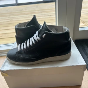 Maison Margiela high tops - Ett par svarta sneakers från Maison Margiela i äkta skinn med vita snören och vit sula.                                             Klassisk high-top modell med diskret logotyp på plösen och ljus insida. Skorna har används sparsamt och är i ett fint skick🤩 