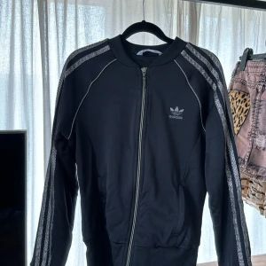 Svart Adidas bomberjacka med dragkedja - Svart bomberjacka från Adidas med klassiska ränder längs ärmarna och logga på bröstet. Jackan har dragkedja framtill, ribbade muddar och sportig look. Perfekt för en avslappnad stil.