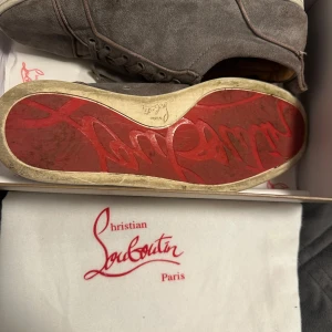 Grå mockasneakers från Christian Louboutin - Säljer ett par grå sneakers från Christian Louboutin i mocka med den ikoniska röda sulan. Skorna har rund tå, platt sula och snörning. Perfekta för dig som vill ha en stilren och lyxig look med en touch av Paris.