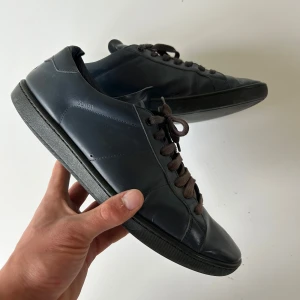 Mörkblå Saint Laurent sneakers i skinn - Snygga mörkblå sneakers från Saint Laurent i slimmad modell. Skorna är tillverkade i skinn med svarta gummisulor och bruna snören. Diskret logga på sidan och häl, samt präglad text på plösen. Perfekta för dig som gillar stilrena och exklusiva sneakers. Hul i sulan som visas. Skriv för mer information🙌