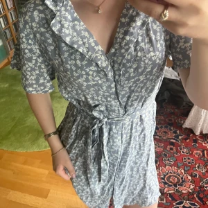 Blommig ljusblå omlottklänning - Supersöt ljusblå kortklänning med vitt blommönster och volangkant vid halsen från Nelly. Omlottmodell med knyt i midjan och korta ärmar. Perfekt för sommarens alla tillfällen! 