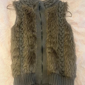 Cool grå/brun stickad pälsväst - Mysig väst från RT i storlek S med stickade partier och flätmönster längs sidorna. Framsidan har mjuka, lurviga pälsdetaljer och västen stängs med dragkedja. Perfekt lager-på-lager-plagg för kyliga dagar. Relativt liten i storleken, passar xxs-s. Inte äkta päls vad jag kan se.
