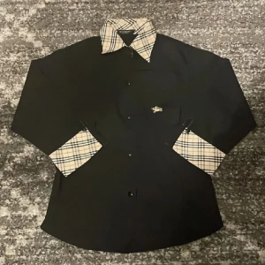 BYTER! - Säljer en svart skjorta från Burberry med klassiska beige-rutiga detaljer på krage och ärmslut. Skjortan har knappar framtill och en liten broderad logga på bröstet. Perfekt för dig som gillar stilrena plagg med en twist.
