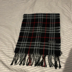 Rutig svart halsduk från Burberrys i ull - Snygg rutig halsduk från Burberrys i 100% lammull. Svart bas med grå och röda rutor, klassisk design och fransar i ändarna. Perfekt accessoar för att lyfta din outfit med en tidlös brittisk vibe.