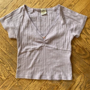 Ljuslila ribbad topp från Urban Outfitters - Supersöt ljuslila topp från Urban Outfitters med v-ringning och liten rosett med blomdetalj framtill. Toppen är ribbad med korta ärmar och har en feminin spetskantp vid halsen. Om blomman skulle vara ett problem är den lätt att klippa bort. Toppen funkar till allt och är så så fin på alla sett.Har använt en gång och säljer så nån annan kan få mer användning för den än jag!