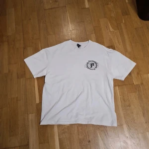 Vit oversized t-shirt från Black Squad med tryck - Säljer en vit oversized t-shirt från Black Squad i storlek XS, passar dock som M eftersom att tröjan är oversized. T-shirten har ett stort tryck med ros och texten 'Paranoid Society' på ryggen och ett mindre tryck på bröstet. Perfekt för dig som gillar streetwear vibes.