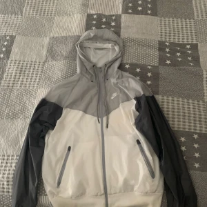 Nike vindjacka i grått och vitt - Nike vindjacka med huva i färgerna ljusgrå, mörkgrå och vit. Jackan har dragkedja framtill, två fickor med dragkedja och justerbar huva. Perfekt för blåsiga dagar och sportiga outfits.☺️☺️