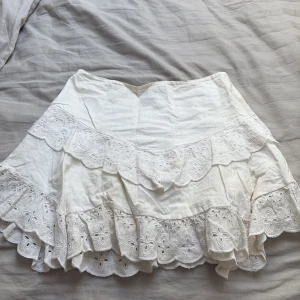 Vit volangkjol med broderade detaljer - Supersöt vit kjol med flera lager volanger och broderade spetsdetaljer. Kjolen har en kort längd och är perfekt för varma dagar. Den har en romantisk och bohemisk känsla med fina blommiga broderier på volangerna. 