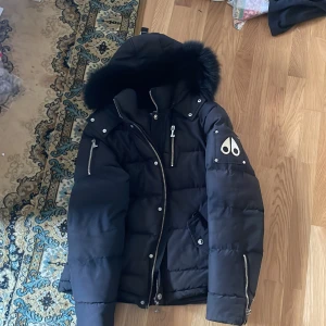 Svart dunjacka med pälskant från Moose Knuckles - Säljer en svart dunjacka från Moose Knuckles med huva och pälskant. Jackan har dragkedja, flera fickor med dragkedjor och logga på ärmen. Perfekt för kalla dagar och riktigt snygg design.de enda är att jag låna ut den och drag kedjan strular lite men den funkar och jag brukar inte använda den jag brukar använda knapparna