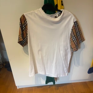 Vit t-shirt med rutiga ärmar från Burberry - Säljer en vit t-shirt från Burberry. Finns ingen skada på plagget då det har användits till få tillfällen. Perfekt att ha till sommaren