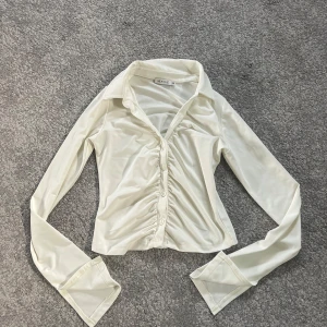 Vit långärmad topp från NLY ONE - Snygg vit skjorta från NLY ONE i storlek XS. Skjortan har lång ärm, klassisk krage och knappar framtill. Den är figurnära med rynkade detaljer längs knappraden och har en lätt glansig finish som ger en trendig look.