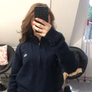 Mörkblå stickad zip-jacka Lacoste - Mörkblå stickad jacka från Lacoste med dragkedja framtill och klassisk krokodil-logga på bröstet. Jackan har lång ärm, ribbade muddar och en clean, sportig look. Perfekt för dig som gillar stilren och enkel design.