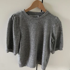 Grå puffärm tröja från 157 - Supermjuk grå tröja från 157 med puffiga trekvartsärmar och rund halsringning. Perfekt för dig som gillar en enkel men ändå trendig look. Materialet känns som bomull och polyester, vilket gör den både bekväm och lätt att bära.
