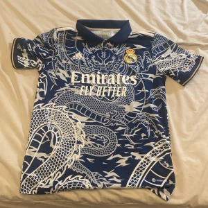 Real Madrid x Adidas blå draktröja S - Säljer en blå Real Madrid fotbollströja från Adidas i storlek S. Tröjan har ett fett drakmönster i vitt över hela plagget, klassisk krage och korta ärmar. Broderad Real Madrid-logga och Adidas-märke på bröstet. Materialet är lätt och andas, perfekt för match eller träning. Högaste priset = 300kr. Lägsta priset = 150