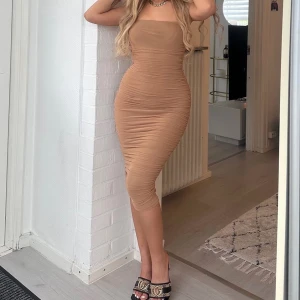 Beige tight klänning med rynkade detaljer - Säljer en beige, tight klänning med rynkade detaljer och ärmlös design. Klänningen har en rak, fyrkantig urringning och är figurnära med midilängd. Perfekt för dig som gillar en stilren och trendig look.