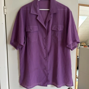 Lila skjorta med fickor och knappar - Säljer en lila skjorta med kort ärm och två bröstfickor med lock och matchande tygklädda knappar. Skjortan har en klassisk krage och är tillverkad i ett lätt, strukturerat tyg som känns luftigt. Perfekt för dig som gillar färg och vill sticka ut lite extra.