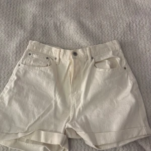 Vita jeansshorts från 157, storlek L - Snygga vita jeansshorts från 157 med klassisk femficksmodell och uppvikta benslut. Hög midja och silverfärgade nitar ger en clean look. Perfekta för varma dagar och lätta att matcha med allt. Materialet är kraftig bomull som känns stabilt och håller formen.