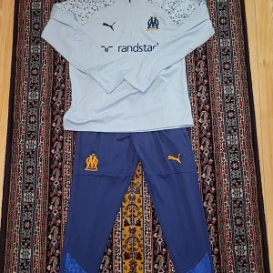 Marseille Tracksuit  - Snygg långärmad ljusblå fotbollströja från Puma för Olympique de Marseille. Tröjan har vita och blå detaljer, diskret mönster på axlarna och klubbmärke samt Puma-logga på bröstet. Materialet är lätt och andas, perfekt för träning eller match.