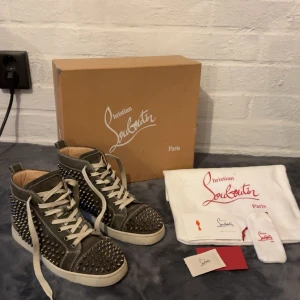 Mörkgröna Christian Louboutin sneakers med nitar - Snygga höga sneakers från Christian Louboutin i olivgrön mocka med silvriga nitar över hela skon. Vita snören, vit sula och klassisk röd undersida. Märkeslogga på sidan och cool kontrast med vita sömmar. Perfekta för dig som vill sticka ut.