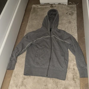 Grå hoodie med dragkedja - Säljer en grå hoodie med hel dragkedja och huva. Tröjan har två stora fickor framtill och en tunn vit linje som detalj längs ärmar och bröst. Perfekt för en avslappnad stil. Det är storlek S