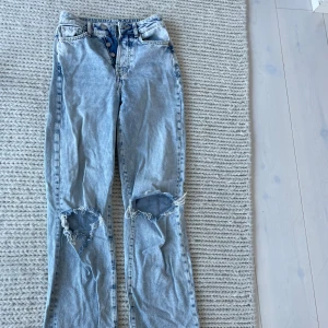 Ljusblå slitna jeans med hål på knäna - Säljer ett par ljusblå jeans med raka ben och stora hål på båda knäna. Jeansen har knappar i gylfen och klassisk femficksdesign. Perfekta för en avslappnad och trendig look.