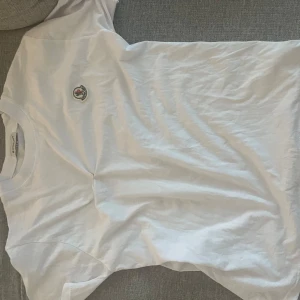 Vit t-shirt från Moncler - Säljer en klassisk vit t-shirt från Moncler med rund hals och Moncler-logga broderad på bröstet. T-shirten är i mjuk bomull och har en enkel, stilren design som passar perfekt till jeans eller shorts.