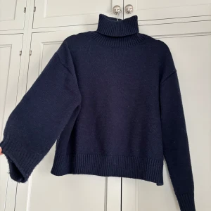 Mörkblå stickad polotröja - Mysig mörkblå stickad polotröja med hög krage och breda ribbade muddar vid ärmslut och nederkant. Tröjan har en loose passform och långa ärmar, perfekt för kyliga dagar. Enkel och stilren design som är lätt att matcha.