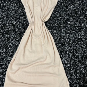 Beige ribbad långklänning med smala axelband - Säljer en beige ribbad långklänning med smala axelband och rak halsringning. Klänningen har en tight passform och är tillverkad i ett mjukt, stretchigt material som sitter snyggt på kroppen. Perfekt för dig som gillar minimalistisk stil. Slit vid benet (inte jätte högt upp)