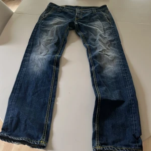 Blå jeans från dondup med slitna detaljer - Säljer ett par blå jeans från Diesel med snygga slitningar och kontrastsömmar i gult och orange. Klassisk femficksmodell med rak passform och coolt D-märke på bakfickan. Perfekta för dig som gillar en avslappnad och streetig stil.