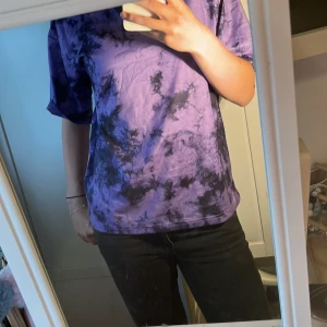 Lila T-shirt - Säljer en lila och svart batikmönstrad t-shirt med loose passform. T-shirten har korta ärmar och är perfekt för dig som gillar en avslappnad och trendig stil. Materialet känns mjukt och skönt mot huden.