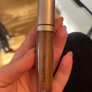 bareMinerals Lipgloss Adventure - Snyggt läppglans från bareMinerals i nyansen Adventure. Färgen är en varm, skimrande brons med guldigt glow. Kommer i en stilren tub med silvrigt lock och applikator. Perfekt för dig som vill ha glansiga, fylliga läppar med metallic-effekt.