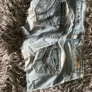 Ljusblå jeansshorts från Tommy Hilfiger - Snygga ljusblå jeansshorts från Tommy Hilfiger med råa kanter och slitna detaljer. Klassisk femficksmodell med logga på bakfickan och patch i midjan. Perfekta för varma dagar och en avslappnad stil.