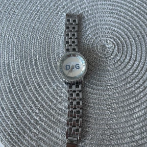 Silverfärgad D&G klocka med stenar - Snygg klocka från D&G i silverfärg med länkarmband i metall. Urtavlan är vit med stora D&G-bokstäver och har glittrande stenar runt boetten. Perfekt accessoar för att lyfta din outfit.