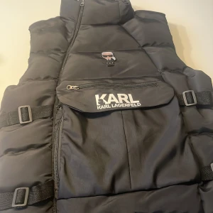 Svart dunväst från Karl Lagerfeld - Svart dunväst från Karl Lagerfeld med cool logga och ikonisk Karl-figur på bröstet. Västen har hög krage, sned dragkedja, justerbara spännen i sidorna och en stor ficka framtill med dragkedja och tryckt logotyp. Perfekt för lager-på-lager-stil.