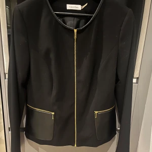 Svart kavajjacka med gulddragkedjor från Calvin Klein - Stilren svart kavajjacka från Calvin Klein med rund halsringning och guldfärgade dragkedjor vid fickorna. Jackan har långa ärmar och detaljer i skinnimitation på fickorna. Perfekt för en dressad look.