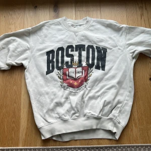 Vit college-tröja Boston Vaillent S - Snygg vit college-tröja från Vaillent i storlek S med Boston University-tryck framtill och broderad logga på ärmen. Tröjan har rund hals, ribbade muddar och är tillverkad i mjuk bomullsmix. Perfekt för en chill och sportig look.