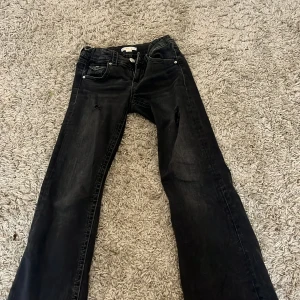 Svarta bootcut jeans från Gina Tricot - Svarta jeans från Gina Tricot med bootcut passform och slitna detaljer på benen. Klassisk femficksmodell med snygga bakfickor med lock och silverfärgade knappar. Jeansen har dragkedja och knapp framtill och är tillverkade i denim.