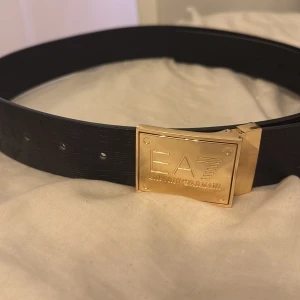 Svart bälte med guldfärgat spänne från EA7 - Snyggt svart bälte från EA7 Emporio Armani med ett stort, fyrkantigt guldfärgat spänne där loggan är ingraverad. Bältet har präglad EA7-text längs hela remmen och är gjort i skinn. Perfekt accessoar för att lyfta din outfit.
