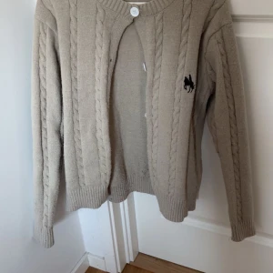 Beige kabelstickad kofta från Ralph Lauren - Kabelstickad beige kofta från Ralph Lauren med rund halsringning och knäppning framtill. Klassisk design med broderad logga på bröstet. Perfekt för lager-på-lager och enkel att matcha med olika outfits.