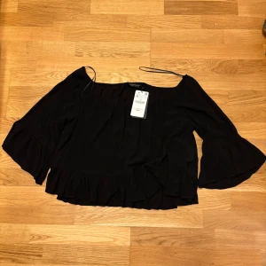 Svart blus med volang från Zara Woman - Säljer en svart blus från Zara Woman med volangdetaljer vid ärmslut och nederkant. Blusen har vida trekvartsärmar och en lös passform, perfekt för en trendig look.