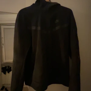 Svart zip-hoodie från Nike - Svart hoodiejacka från Nike med dragkedja framtill och diskret logga på bröstet. Jackan har lång ärm, ficka med dragkedja på ena ärmen och detaljer i form av svarta band över bröstet. Perfekt för en sportig stil.
