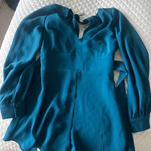 Blå långärmad jumpsuit från River Island - Säljer en stilren blå jumpsuit från River Island med långa ärmar och markerad midja. Den har en snygg v-ringning med knyt i halsen och manschetter med knappdetalj. Perfekt för dig som vill ha en enkel men elegant look 👗