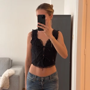 Svart croppad topp med spets och knyt - Säljer en svart croppad topp med spetsdetaljer längs kanterna och två knyt framtill. Toppen har breda axelband och v-ringning, perfekt för sommaren.🩷