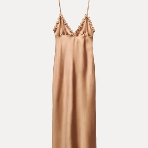 Champagnefärgad långklänning  - Supersnygg långklänning i champagnefärgad satin från Zara. Klänningen har smala axelband, v-ringning med volangdetaljer och en slimmad passform som faller snyggt längs kroppen. Perfekt för dig som vill sticka ut med en elegant och trendig look. Helt oanvänd i stolek xs 