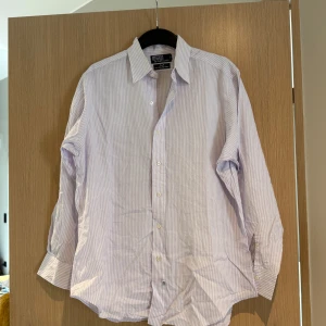 Randig skjorta från Polo Ralph Lauren - Klassisk skjorta från Polo Ralph Lauren i vitt och ljuslila med tunna ränder. Skjortan har lång ärm, knappar framtill och är tillverkad i 100% bomull. Perfekt för dig som gillar en stilren och tidlös look.
