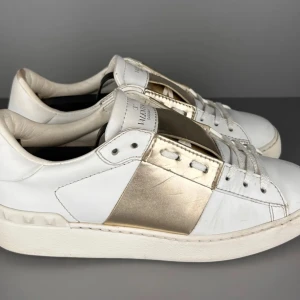 Valentino Garavani vita sneakers med guld - Snygga vita sneakers från Valentino Garavani med bred guldfärgad detalj över mitten. Klassisk låg modell med snörning och rund tå. Skorna är i skinn och har diskreta nitar baktill på sulan för en extra touch.